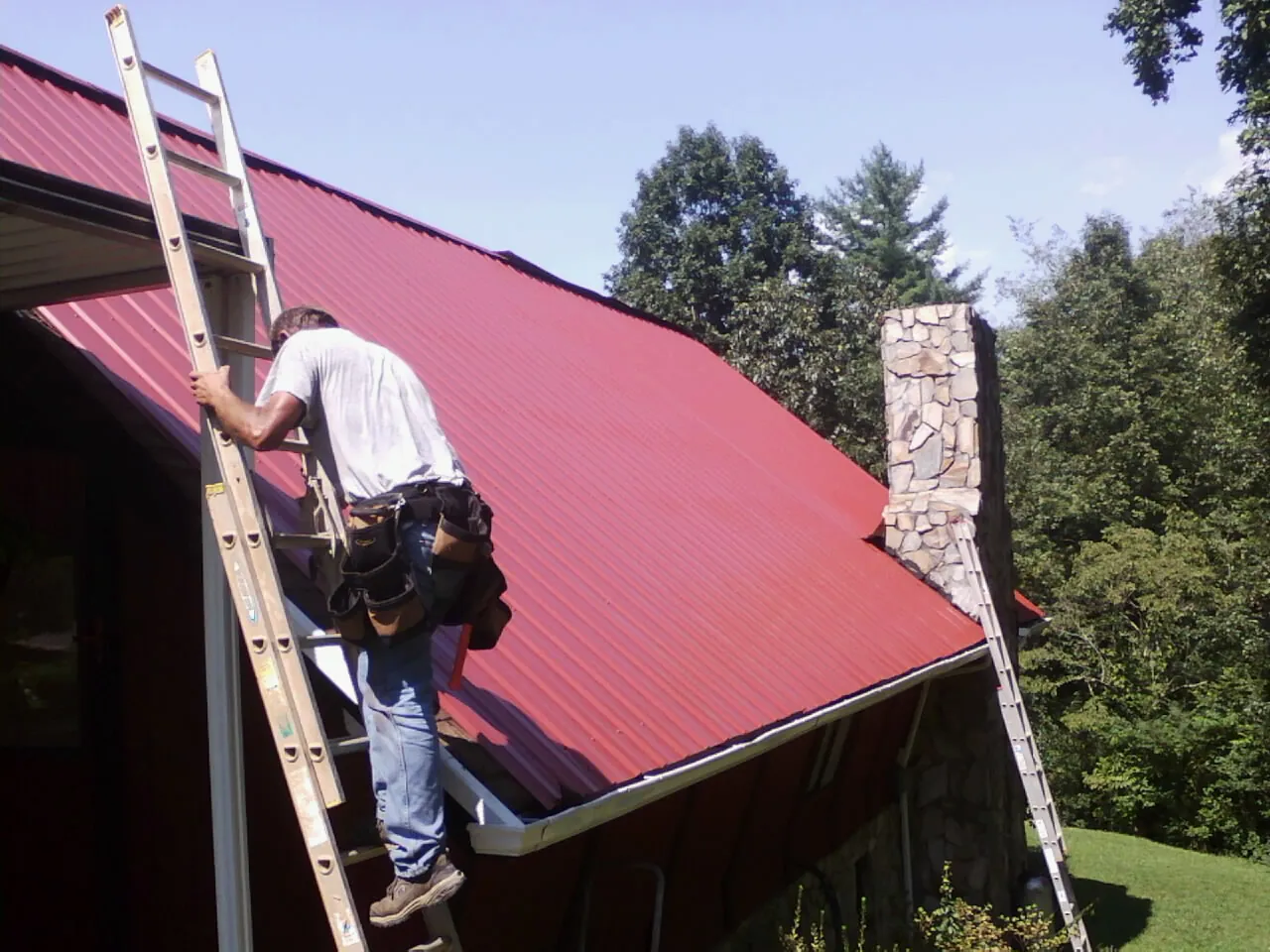 Roofer on a ladder beside a red metal roof and stone chimney — Soffit & Fascia Repair by Roofing Brothers in Middletown
